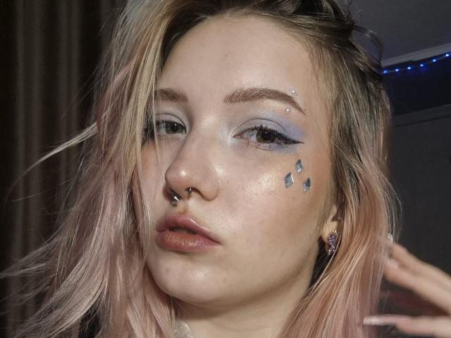 PalmiraEsquinaldo tem cabelo rosa claro, sombra azul, adesivos faciais de diamante, um piercing no nariz e lábios brilhantes. Sua pele está brilhante, com uma expressão séria. Ela está tocando o cabelo..