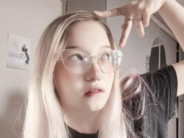 PalmiraEsquinaldo, com pele clara, cabelo loiro-rosa, óculos de grau, piercing no nariz, camisa preta, boca ligeiramente aberta, apontando para a cabeça, anel no dedo..