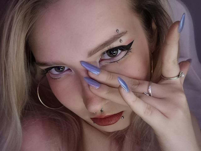 O rosto de PalmiraEsquinaldo, com unhas azuis, delineador, e piercings no nariz e no lábio inferior, cobrindo a boca com a mão, usando um anel de ouro e um brinco de argola..