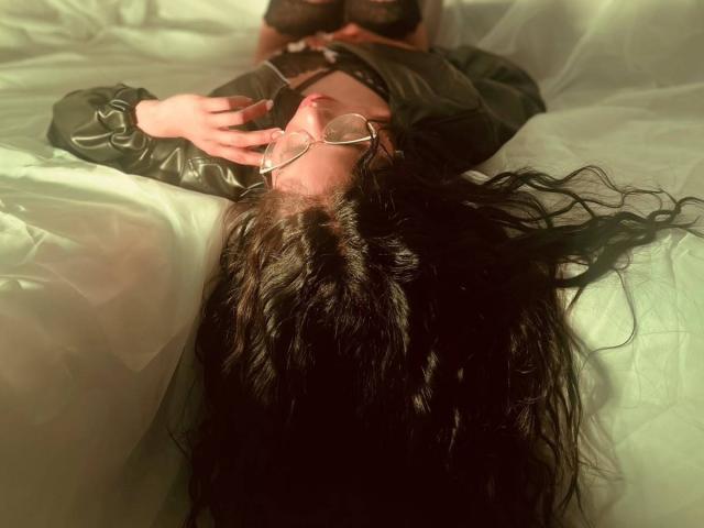 MelannieRousse liegt auf einem Bett, ihr langes, dunkles Haar ist ausgebreitet, sie trägt eine Brille und eine grüne Lederjacke, und ihre Hand befindet sich in der Nähe ihres rot geschminkten Mundes..