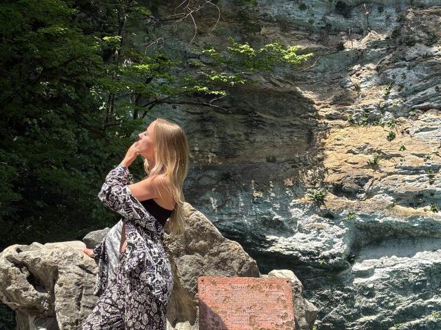AngeloBradac with long blonde hair, wearing a black and white patterned dress, sits on rocky terrain, hand near mouth, looking up with a thoughtful expression.