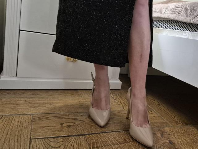 BagiraNoa's legs in a black sparkly skirt, wearing beige pointed high heels, standing on wooden floor. Her pale skin is visible, with a white bedside table in background.