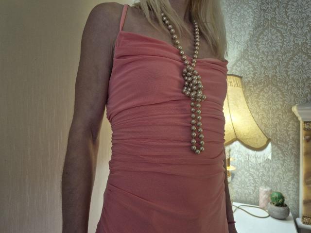 BagiraNoa with blonde hair wearing a ruched pink dress, long pearl necklace, and no visible breasts or pussy. Arm visible, standing near a lit lamp.