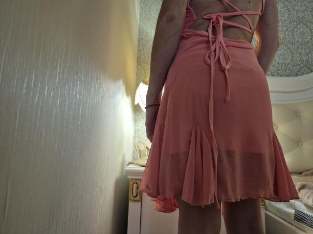 BagiraNoa's pink, see-through, backless dress with lace-up sides and a flared skirt, exposing her ass and lower back, standing in a dimly lit room.