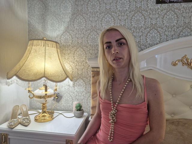 BagiraNoa with blonde hair, pink dress, and pearl necklace, sitting on a white bed, next to a gold lamp and white nightstand with high heels and cactus.