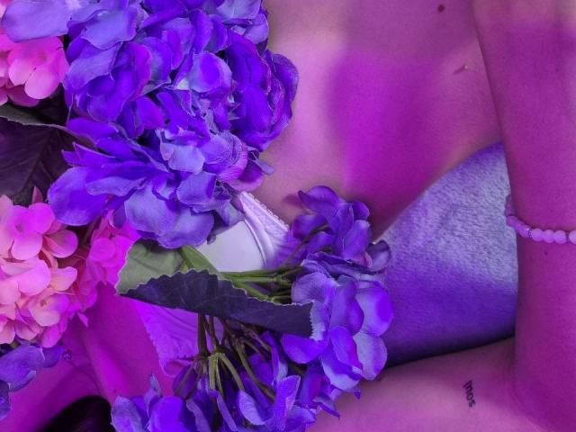 TenerliRize's tits are covered by a white dress with purple and pink flowers on her chest and left arm, wearing a pink bead bracelet.