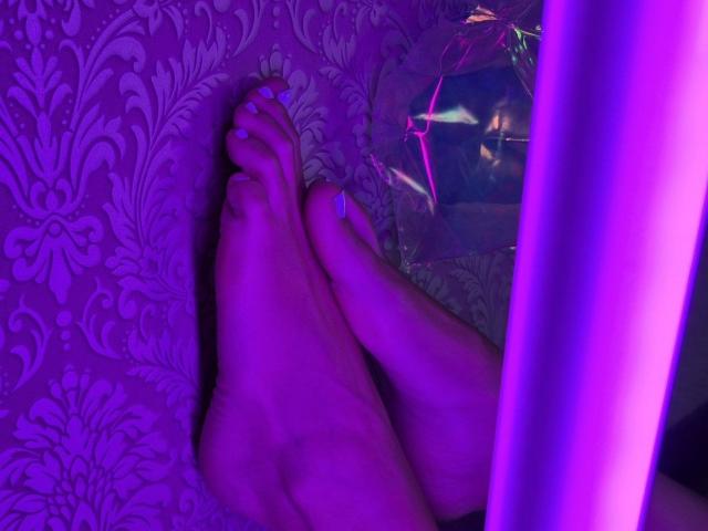 TenerliRize's bare feet, crossed at the ankles, with purple neon lighting. TenerliRize's toes have blue nail polish. A reflective surface is on the right.