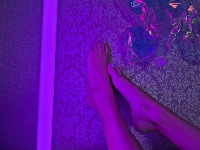 TenerliRize's bare feet crossed on a purple-patterned floor, illuminated by pink and purple neon light, with a shiny, crumpled, colorful object to the right.