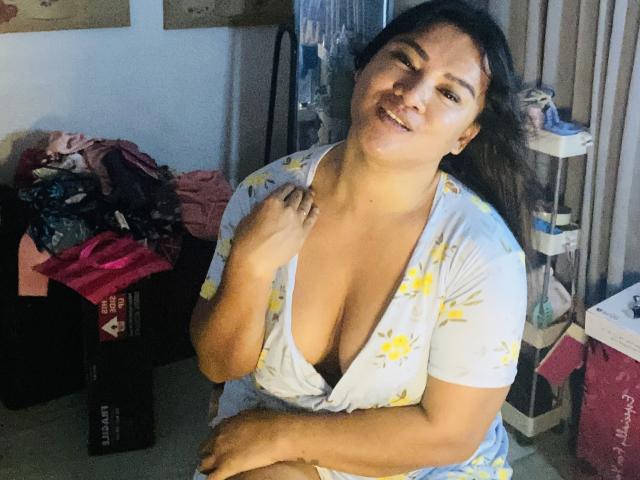 Une jeune femme philippine aux traits doux, à la peau brun clair, aux cheveux foncés et au sourire, portant un haut blanc à fleurs à décolleté, dévoilant sa poitrine, assise dans une pièce en désordre, une main sur la poitrine..