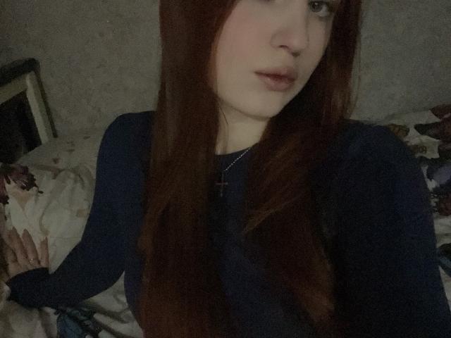 Kattina, con lunghi capelli castani, pelle chiara e occhi azzurri, indossa una camicia scura, guarda verso la fotocamera, ha un naso piccolo, le labbra leggermente dischiuse, si vede una collana, l'illuminazione è fioca..