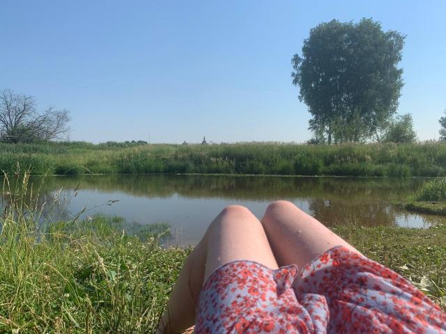 Emerald Crivaro mit roten Shorts mit Blumenmuster, sitzend auf dem Gras an einem Teich mit Spiegelung, blauer Himmel, hohes grünes Gras und ein großer Baum im Hintergrund..