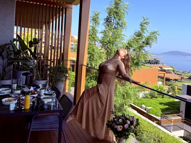 BadAngel, con lunghi capelli biondi, grandi seni e un vestito marrone, è appoggiata alla ringhiera di un balcone, con le mani sui fianchi, e guarda verso l'alto, circondata da piante e con una vista soleggiata..