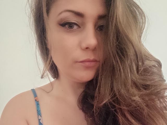 JuliaStormcb with wavy brown hair, blue floral strap, and subtle eyeliner, has a serious expression, showing off her nose and full lips. Her tits are partially visible.