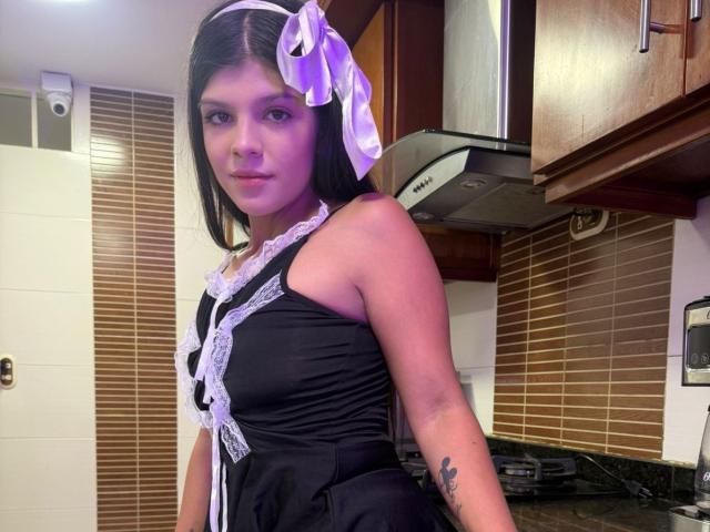 RilleyFoyx mit violett schimmernder Haut, schwarzem Schnürkleid, weißem Band, violettem Schleifchen, kurzen schwarzen Haaren, Tattoo am rechten Arm, stehend in der Küche, Blick zur Kamera, leichtes Lächeln..