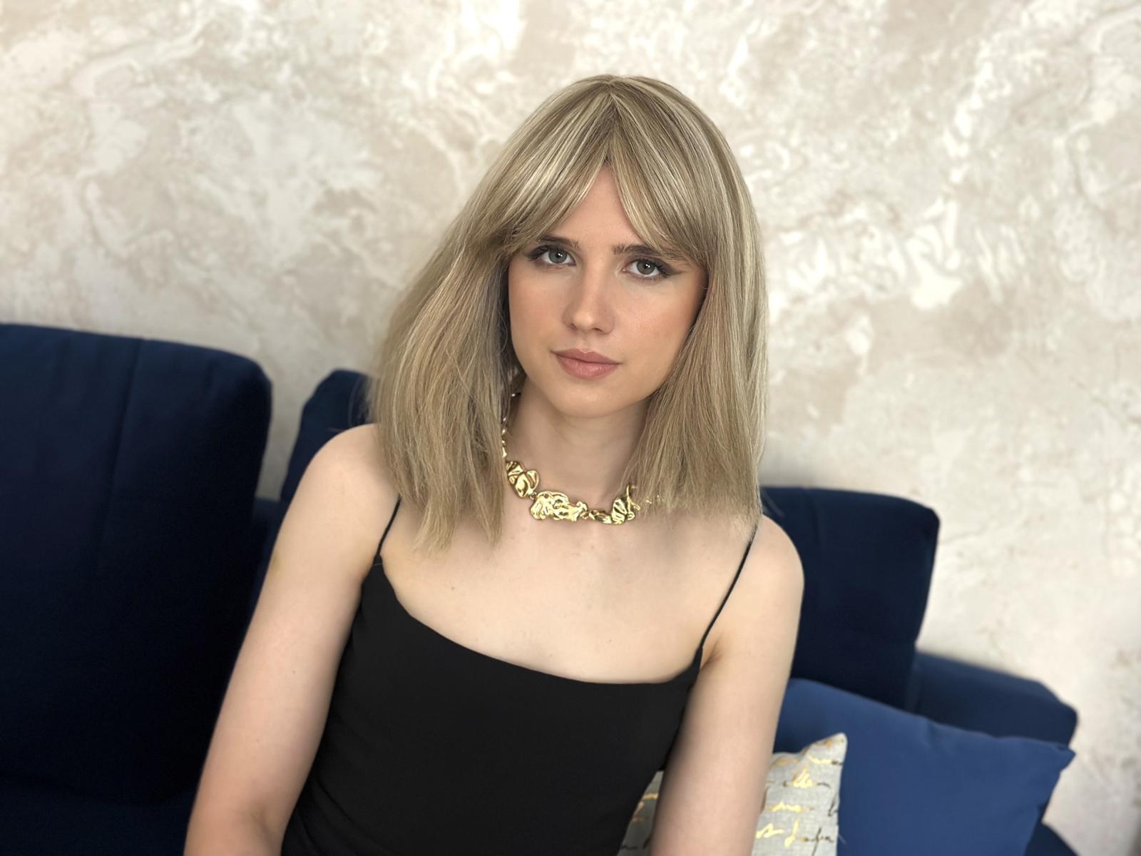 Mia Haris, con cabello rubio, vestida con un top negro de tirantes finos y un collar dorado, sentada en un sofá azul, tiene un rostro lindo y pequeños senos..