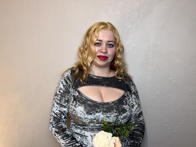 OliviaVerr, una donna bionda di taglia curvy con i capelli ricci, indossa un abito di velluto argentato con un taglio, tiene una rosa, ha il rossetto rosso e un leggero sorriso..