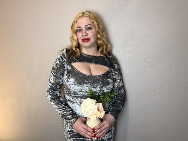 OliviaVerr, con i capelli biondi e ricci, indossa un abito di velluto grigio con dettagli ritagliati, tiene in mano una rosa bianca e del verde, ha il rossetto rosso e sta in piedi di fronte a un muro spoglio..