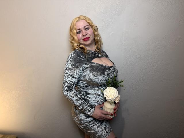 OliviaVerr, bionda con capelli ondulati, indossa un abito di velluto grigio con una scollatura, tiene una rosa bianca, è in piedi contro un muro beige, sorride con il rossetto rosso..