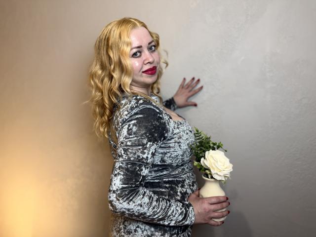 OliviaVerr con capelli biondi ricci, rossetto rosso, vestito grigio a fantasia, che tiene una rosa bianca in un vaso, con una mano appoggiata al muro, che guarda indietro, incinta, leggermente robusta, in piedi..