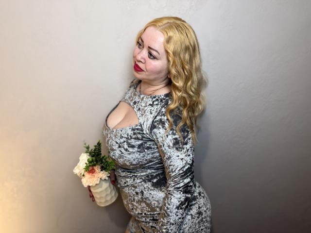 OliviaVerr, con i capelli biondi ondulati, rossetto rosso e un vestito grigio a fantasia, tiene in mano un vaso di fiori, mostrando il suo seno prosperoso e i suoi fianchi formosi..