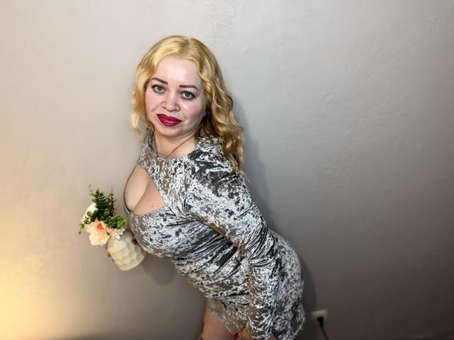 OliviaVerr, con i capelli biondi e ricci, il rossetto rosso e l'abito grigio a fantasia, che mette in mostra il suo seno prosperoso e i fianchi formosi, appoggiata a un muro con un vaso di fiori..