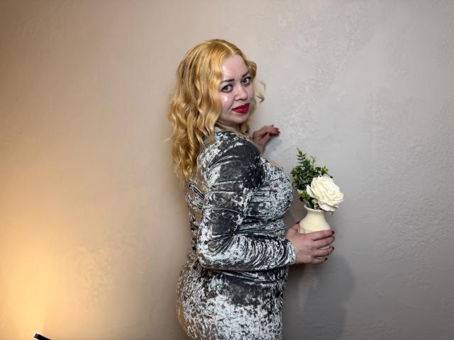 OliviaVerr, con i capelli biondi e ondulati, indossa un abito grigio in velluto aderente, tiene in mano un vaso bianco con una rosa, è in piedi contro un muro e guarda oltre la spalla, con il rossetto rosso..