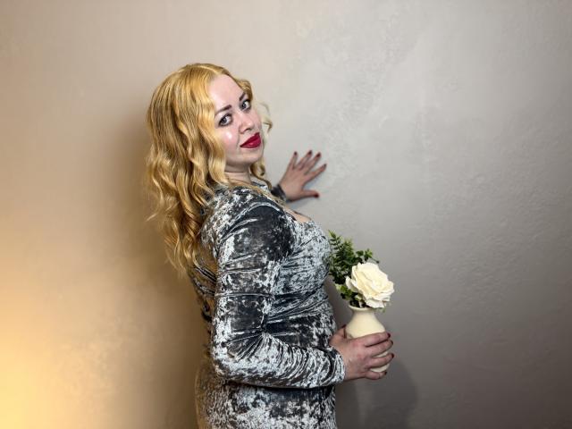 OliviaVerr con capelli biondi ondulati, rossetto rosso, vestito di velluto grigio, che tiene una rosa bianca, con una mano appoggiata al muro, che guarda indietro, mostrando il sedere, in piedi, all'interno, illuminazione soffusa..
