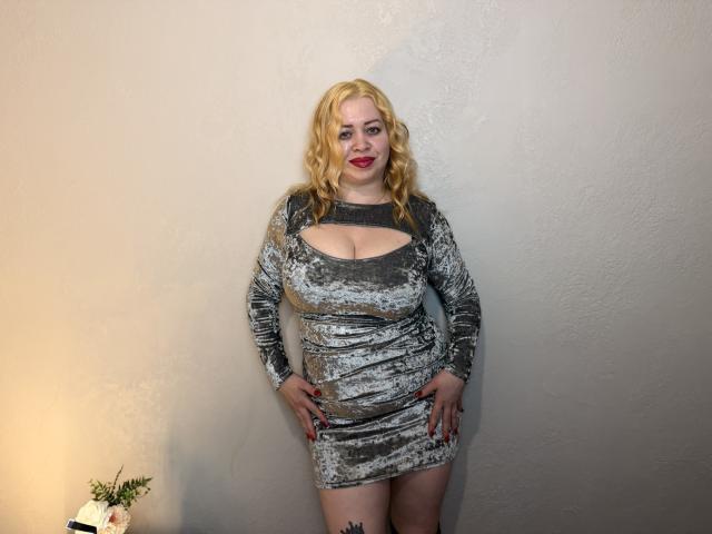 OliviaVerr ha i capelli biondi e ricci, rossetto rosso e indossa un abito di velluto argentato con un taglio che mette in mostra il suo seno prosperoso. È in piedi contro un muro bianco, con le mani sui fianchi..