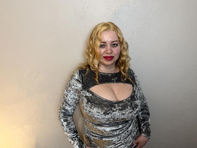 OliviaVerr, con i capelli biondi e ricci, rossetto rosso e un abito di velluto grigio con aperture che mostrano il suo seno prosperoso, in piedi contro un muro spoglio..