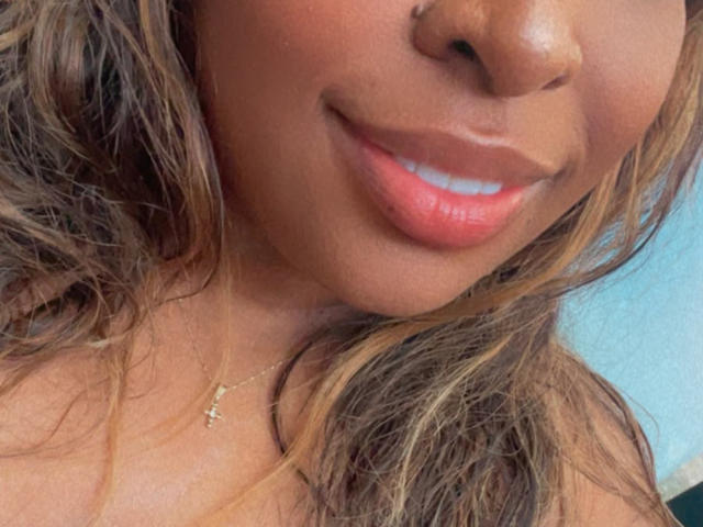 NatyLm's face, showing her brown nose, shiny pink lips, and wavy blonde-brown hair with a small gold necklace. Her smile is subtle, and her skin is dark.