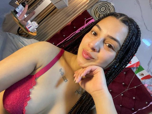 LonxEva with braided hair, pink lace bra, pouty lips, hand on cheek, tattoo under boob, in bedroom. Boobs visible, pink bra straps, soft bedroom light.