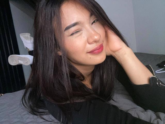 SashaHans, com cabelo preto, olhos fechados e lábios rosados, sorrindo levemente, com a bochecha apoiada na mão, vestindo uma blusa preta, com meias brancas ao fundo..