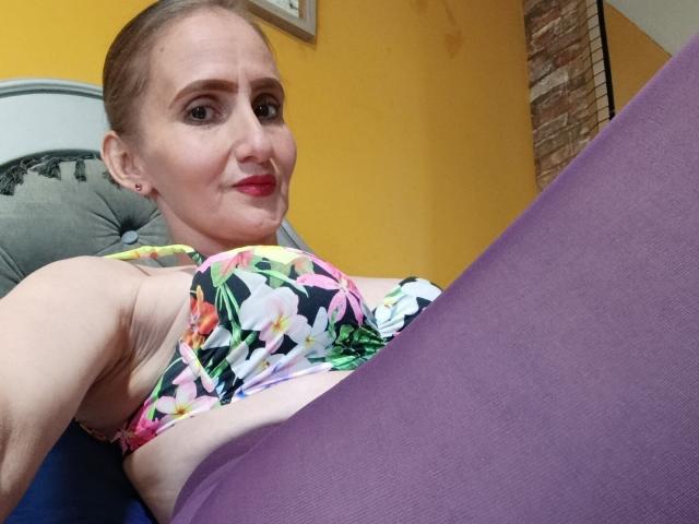 RubiAnderson, con i capelli castano chiaro, rossetto rosso, top da bikini con fiori e leggings viola, è sdraiata, mostrando il seno e l'ascella. Sorride leggermente, guardando la fotocamera..