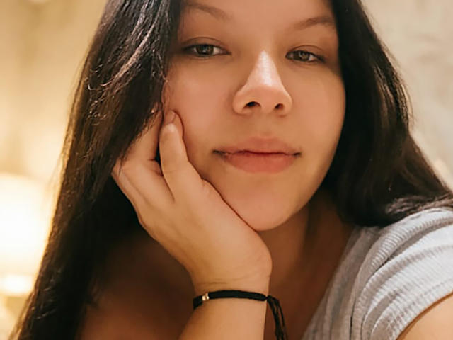 Delilath tiene el cabello largo y oscuro, ojos verdes y una ligera sonrisa. Está apoyando su mejilla en la mano, mostrando una pulsera negra en su muñeca..