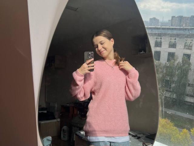 CaroylnPazmino, con una sonrisa encantadora, un suéter rosa y jeans, está tomando una selfie frente al espejo, mostrando sus pequeños pechos y glúteos, en una habitación iluminada por el sol, sosteniendo un teléfono..