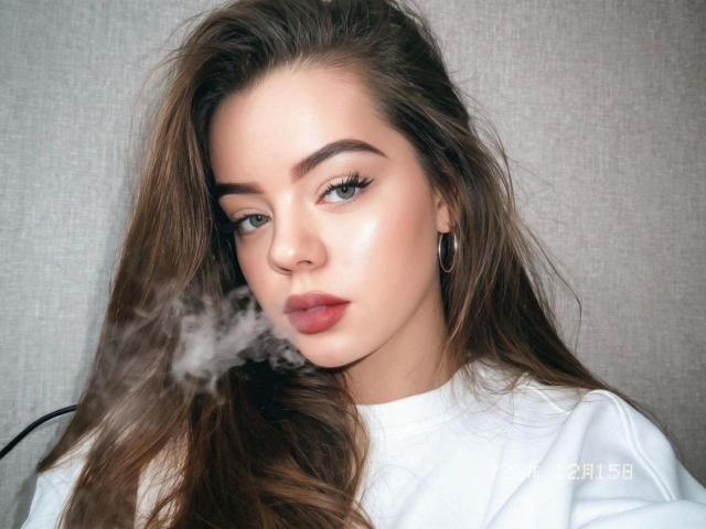 AnasteishaXXX, attraente con lunghi capelli castani, labbra piene e orecchini a cerchio, sta fumando una piccola nuvola di fumo bianco, indossa una camicia bianca, ha gli occhi azzurri e un viso liscio..