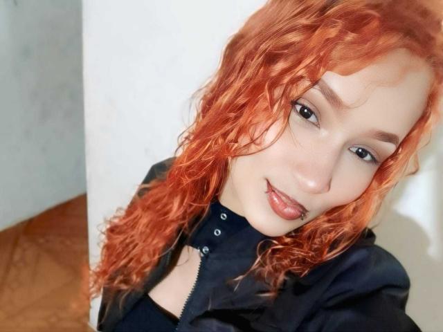 NaomyeLm, com cabelo ruivo ondulado, pele clara e olhos escuros, língua ligeiramente para fora, vestindo um colar preto e jaqueta, sorrindo para a câmera. Seios parcialmente visíveis..