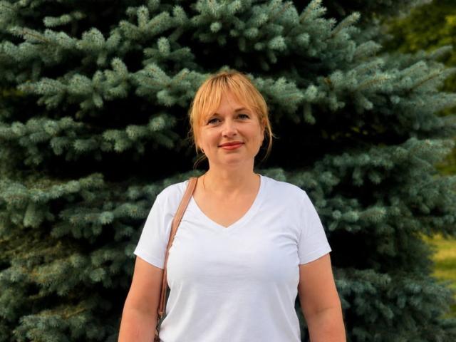 Kira, een lachende, blonde vrouw met kort haar, een witte V-hals top, lichte huid, kleine borsten, een bruine schoudertas, staand voor een grote groene dennenboom, lachend..