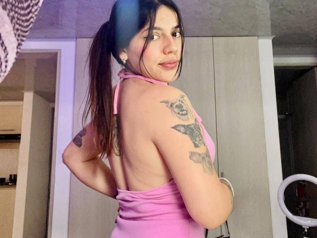 SophiaMarx with pigtails, pink halter top, showing back tattoos, looking over shoulder, smiling, pink lips, ring light in background, white cabinets, arm tattoos, medium skin, cute.
