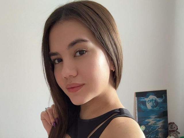 Santina Schaumberg, con cabello castaño largo, piel clara, nariz pequeña, una sonrisa sutil, vestida con una camiseta negra sin mangas, mirando a la cámara, mano derecha con un anillo, una pintura en el fondo..