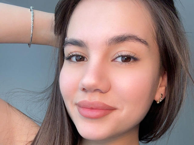 El rostro de Santina Schaumberg, ojos marrones, cabello largo y oscuro, sonrisa sutil, labios rosados, piel clara, cejas finas, pulsera de plata en el brazo izquierdo, pequeños aretes de oro, ligero rubor en las mejillas..