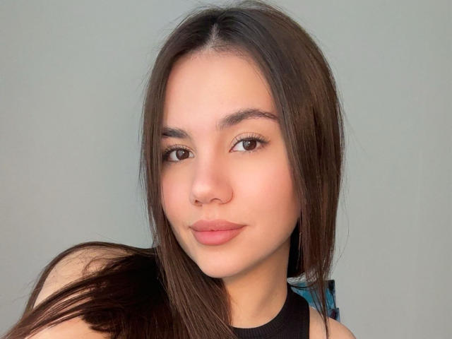 Santina Schaumberg tem cabelo castanho liso, olhos castanhos e lábios rosados com um sorriso discreto, vestindo uma blusa preta sem mangas. Sua pele é clara e lisa..