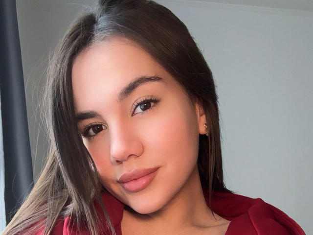 Santina Schaumberg ha gli occhi marroni, i capelli lunghi e scuri, un nasino carino con un trucco leggero, indossa una camicia rossa e un piccolo orecchino dorato all'orecchio sinistro..