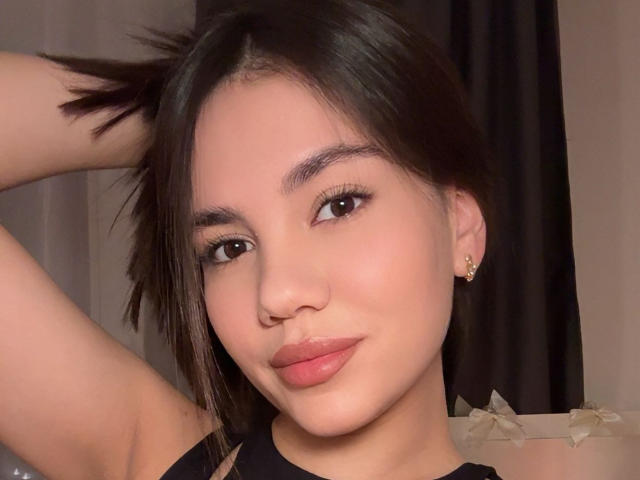 Santina Schaumberg, met donkerbruin haar, lichte huid en een kleine neus, lachend met roze lippen, draagt een zwarte top en oorbellen, heeft haar arm omhoog en draagt subtiele make-up, met zacht licht op haar gezicht..