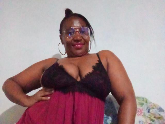 CavyQueenForU, con piel oscura, gafas y lápiz labial rosa, vestida con un camisón negro de encaje y un top rojo transparente, mostrando senos grandes, sonriendo, con las manos en las caderas, el pelo recogido en un moño..