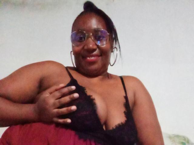 CavyQueenForU, eine dunkelhäutige, kurvige Frau mit Brille, roten Augen, einem schwarzen Spitzenoberteil, die ihre großen Brüste hält, lächelnd, mit schwarzen Haaren im Dutt, trägt Creolen, im schwachen Licht..