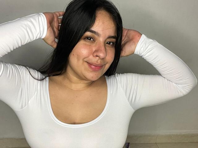 SammyTitss, eine lateinamerikanische Frau mit mittlerem Hautton und schwarzen Haaren, trägt ein weißes Langarmshirt, lächelt und hat die Hände hinter dem Kopf, wobei sie kleine, wohlgeformte Brüste zeigt..
