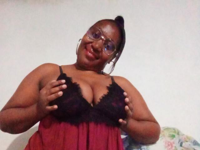 CavyQueenForU con la pelle scura, gli occhiali e un reggiseno nero in pizzo, che tiene le sue grandi, rotonde e piene tette, sorridendo, indossando un baby doll rosso trasparente e una coda alta..