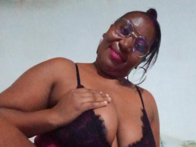 CavyQueenForU, uma mulher de pele escura com óculos, está usando um sutiã de renda preto, segurando seus seios grandes, sorrindo e tem o cabelo preso em um rabo de cavalo..