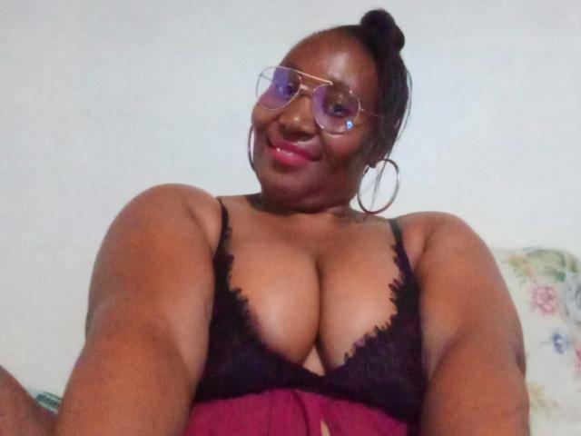 CavyQueenForU, com pele escura, óculos e um coque, vestindo um sutiã de renda preto, mostrando seios grandes, sorrindo, batom rosa e brincos de argola, tirando uma selfie..